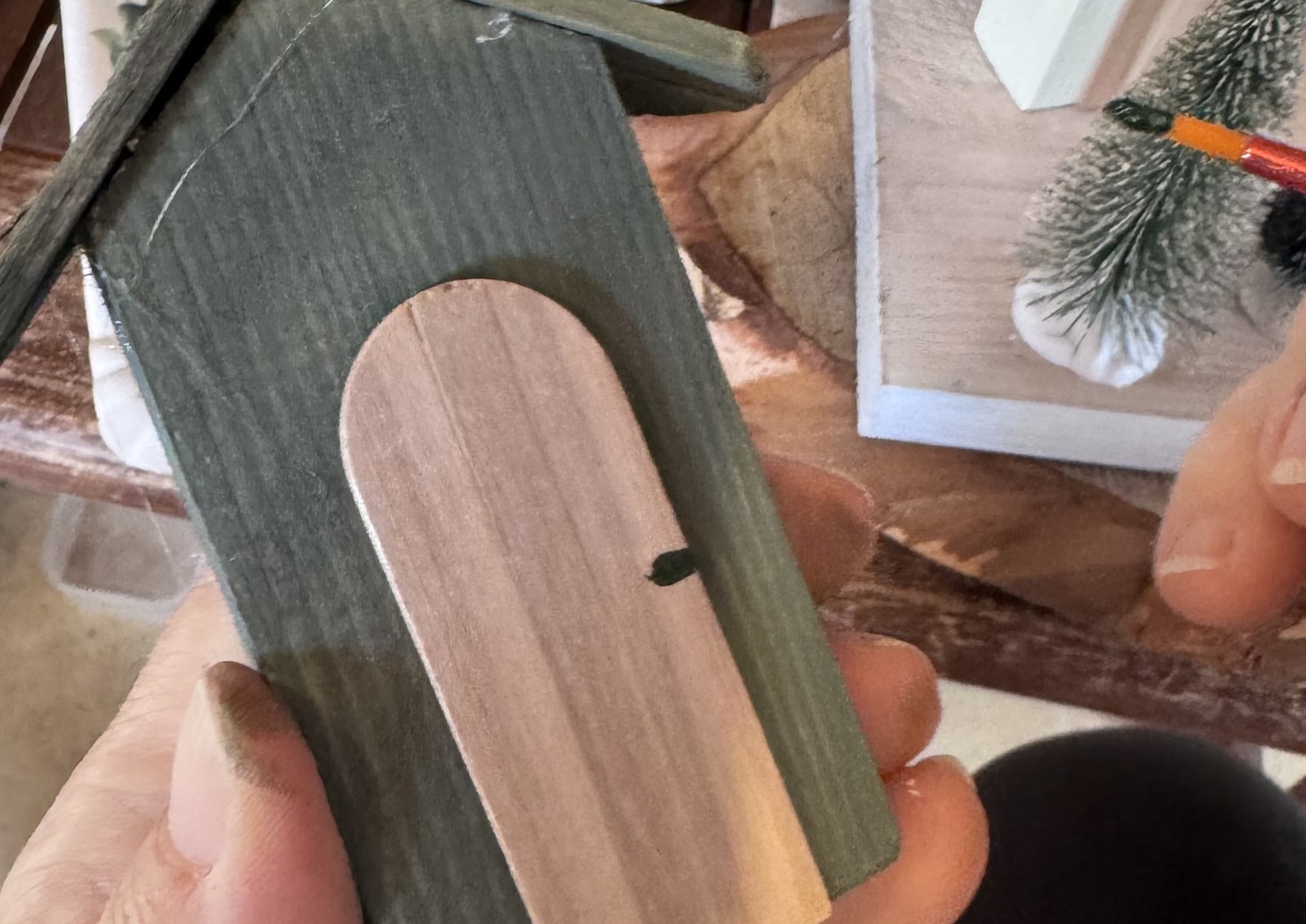 Fotó: egy kéz tart egy zölden festett mini fa házikót, a másik egy havas mini fenyőt fest; az asztalon festéktálca, ecsetek és egy natúr fa házikó.