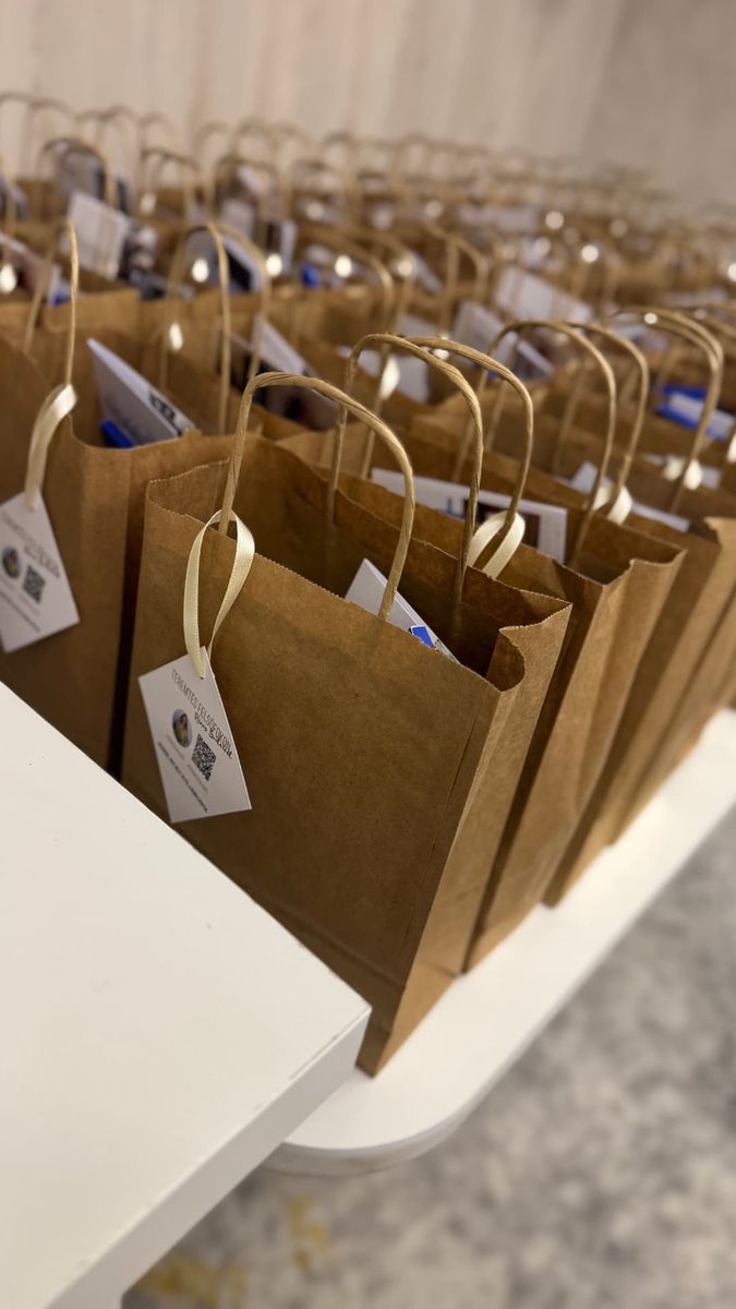 Fotó: barna kraft papírtáskák sora egy fehér asztalon, bennük kiadványok, a fülükre fehér szalaggal kötött kártyákkal.