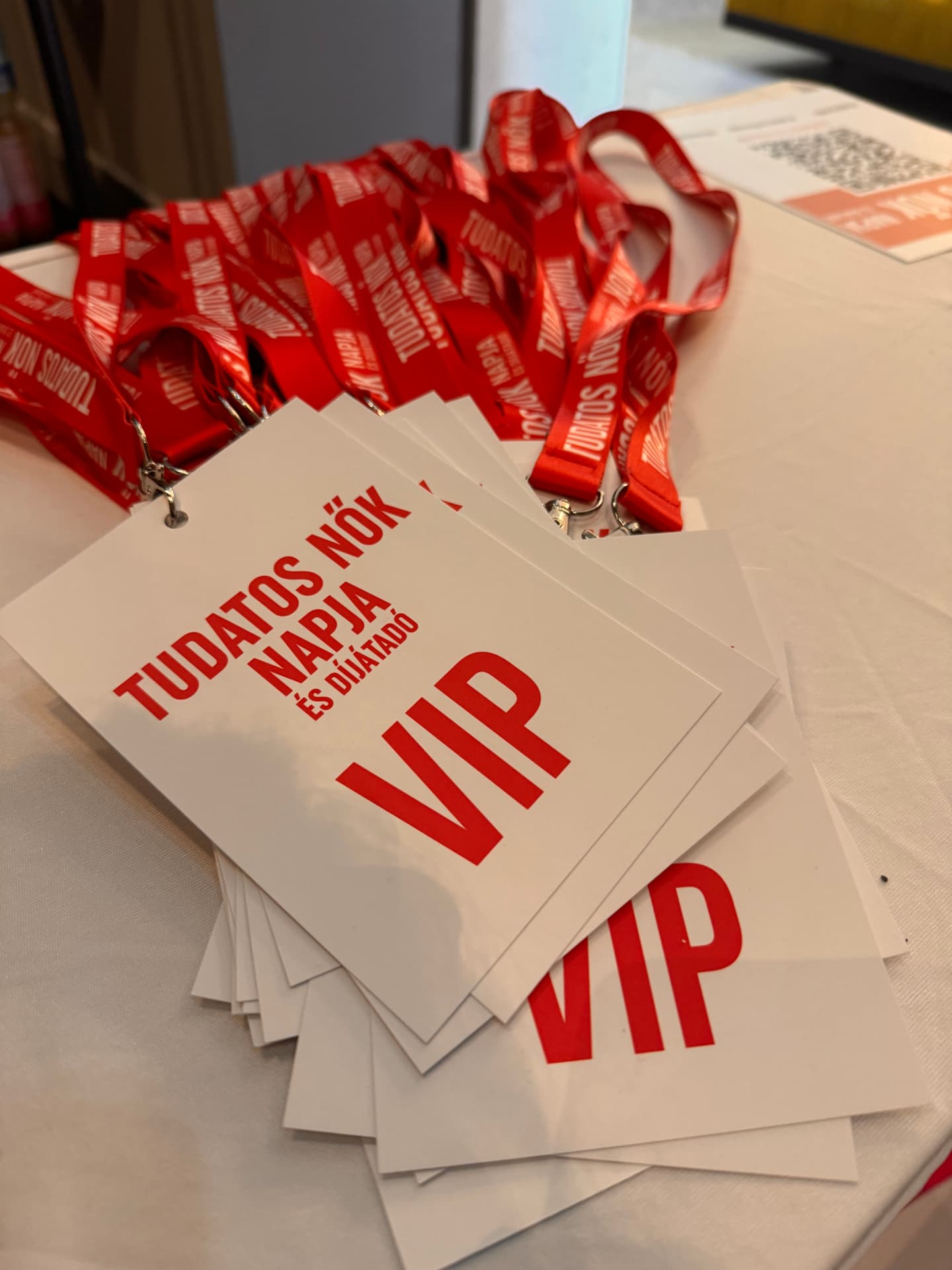 Fotó: fehér, „VIP” feliratú belépőkártyák halma egy fehér asztalon, a rajtuk lévő piros, „Tudatos Nők Napja és Díjátadó” feliratú nyakszalagokkal.