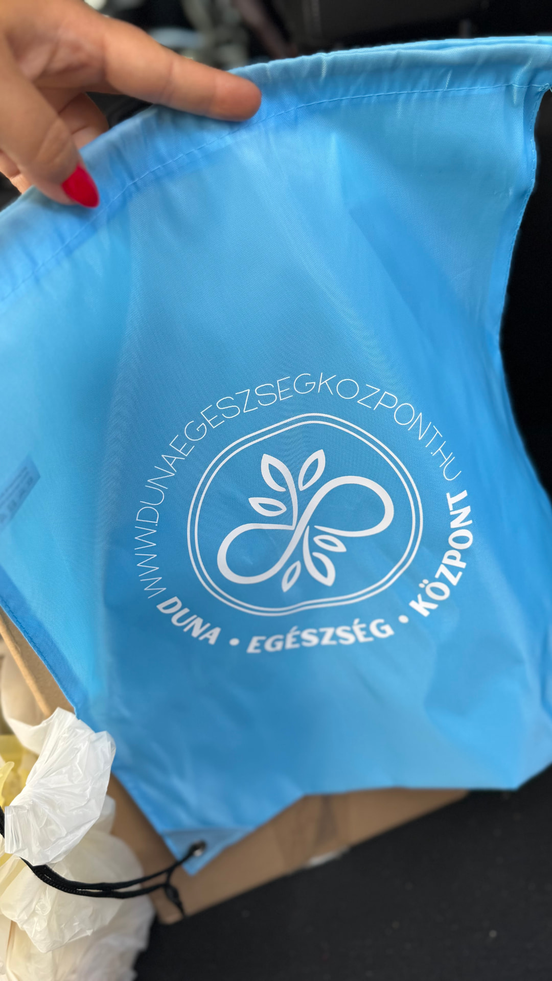 Fotó: egy kézben tartott világoskék, húzózsinóros táska a „Duna Egészség Központ” logójával és webcímével.