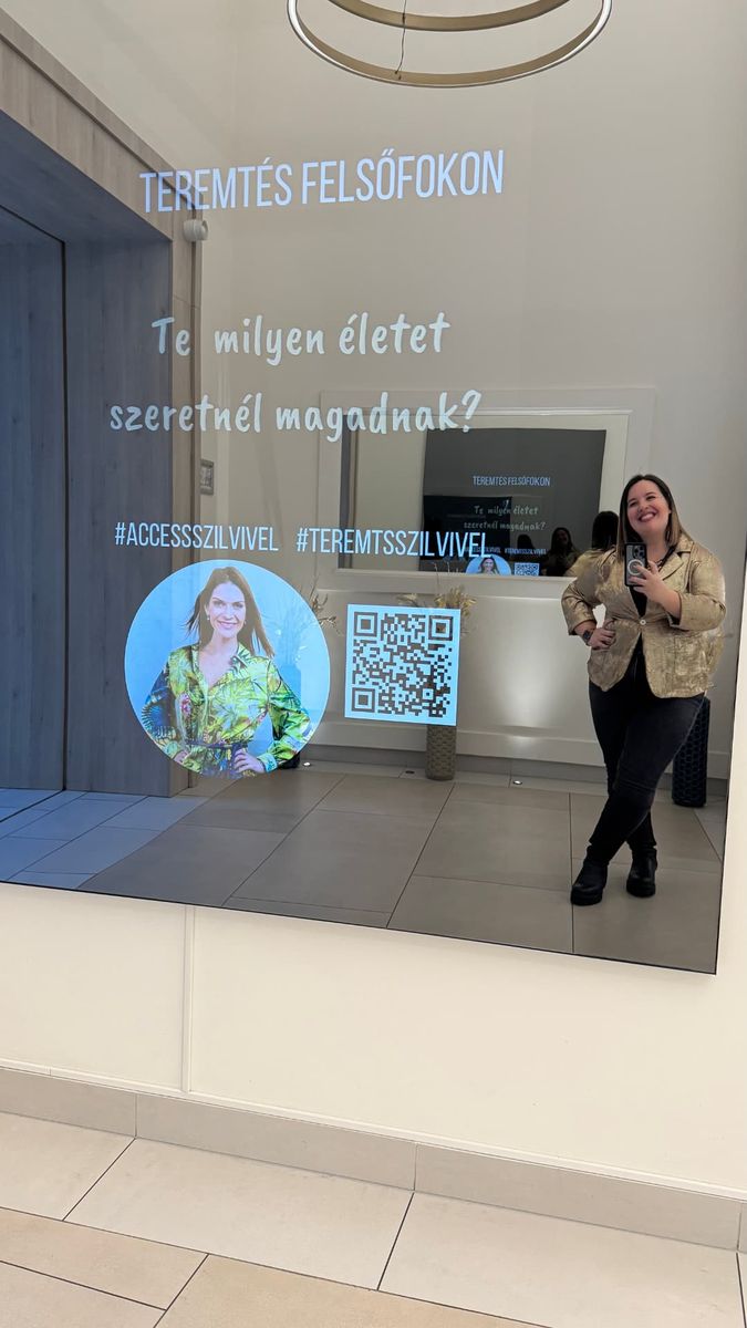 Fotó: arany zakót viselő, mosolygó nő tükörszelfit készít egy modern épület előterében, a „Teremtés Felsőfokon" feliratú inspirációs fali tükör előtt.